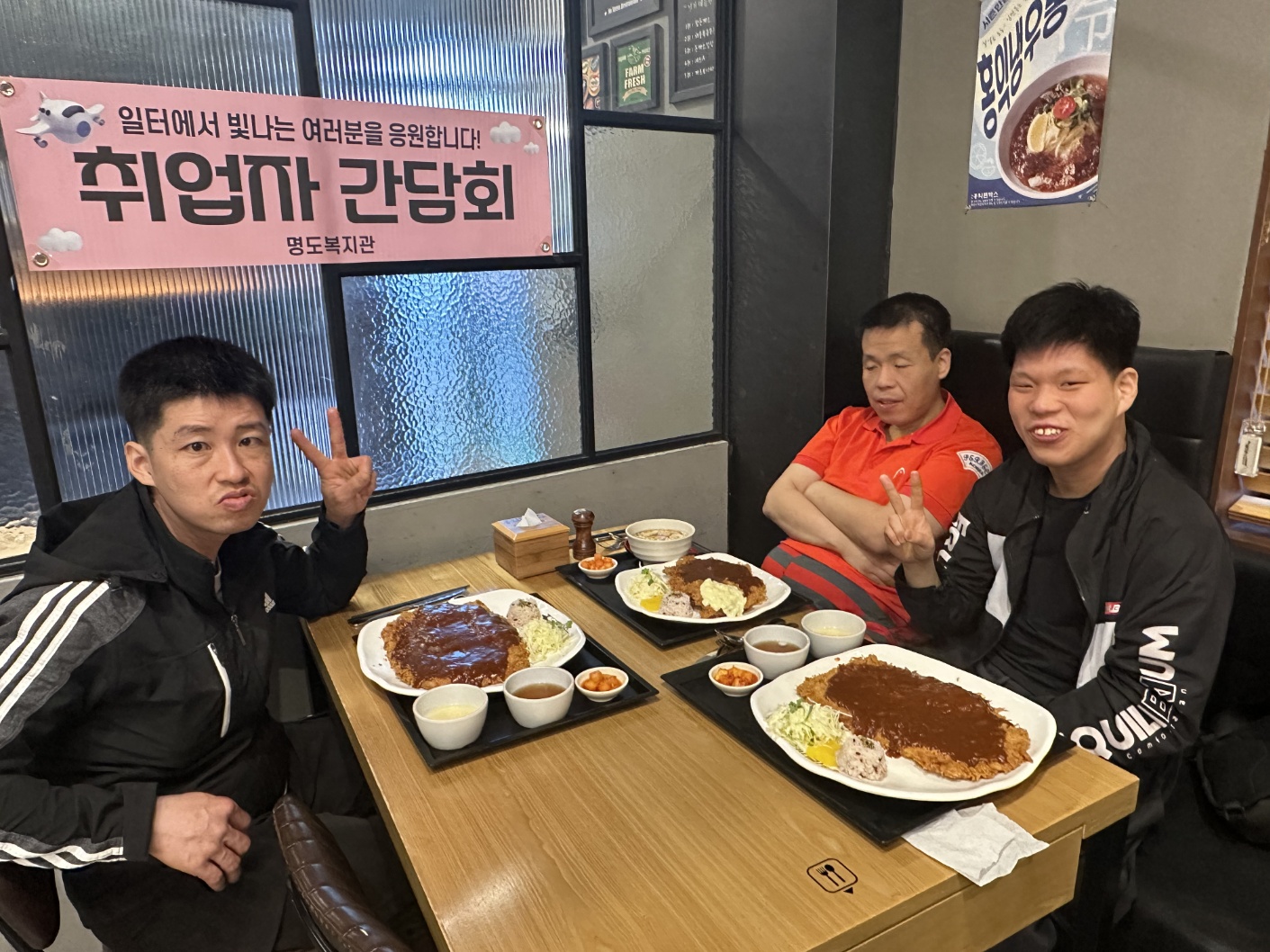 취업자 간담회라고 적힌 현수막이 벽에 걸려 있고, 주문한 돈가스가 테이블에 놓인 채로 취업자 3명이 함께 찍은 기념사진입니다. 