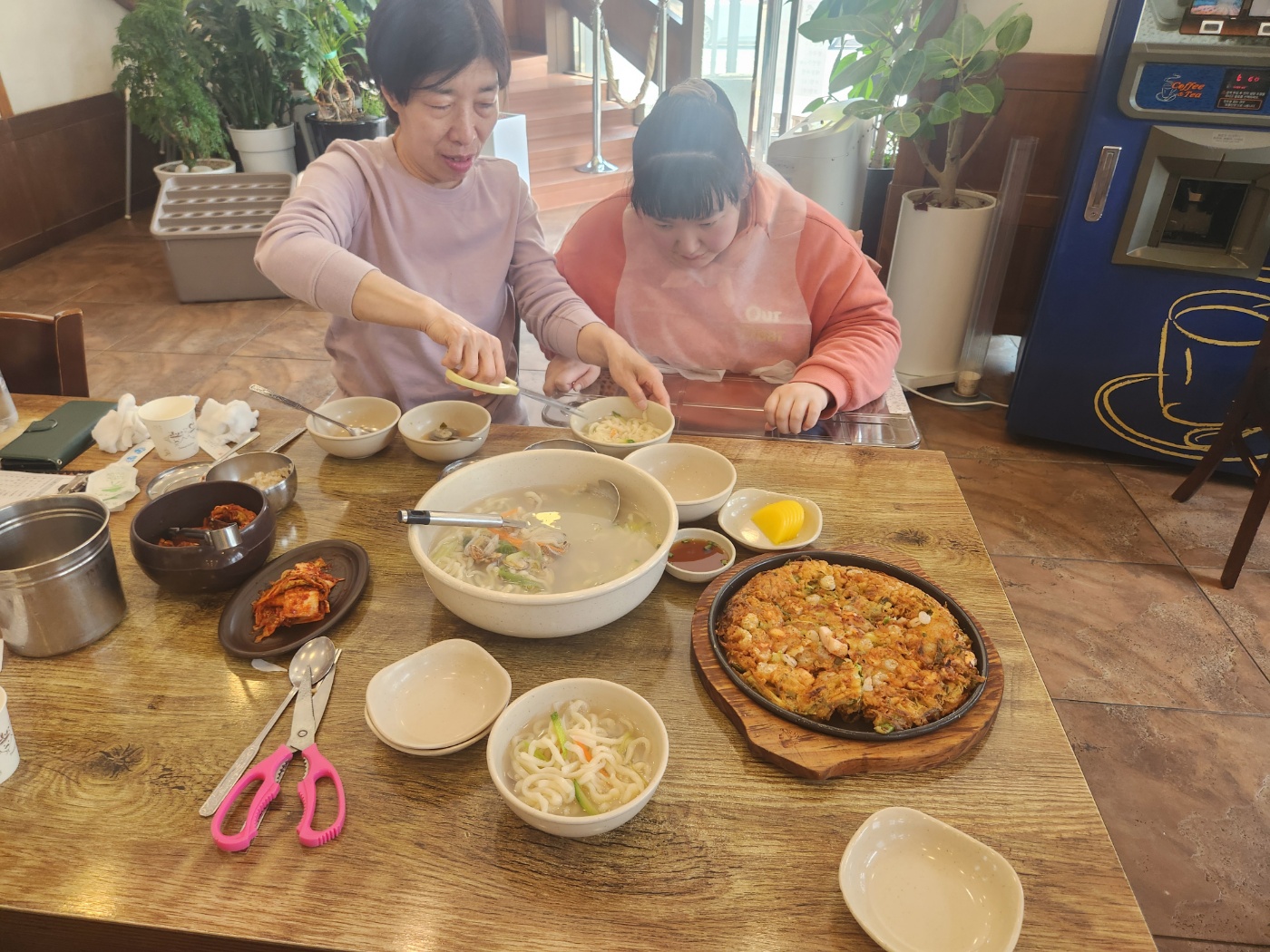 식당에서의 사진으로 주문한 칼국수와 파전이 나와 상에 올려져 있고 활동지원사가 이용인과 나란히 앉아 칼국수를 잘게 잘라주고 있는 모습의 사진입니다. 