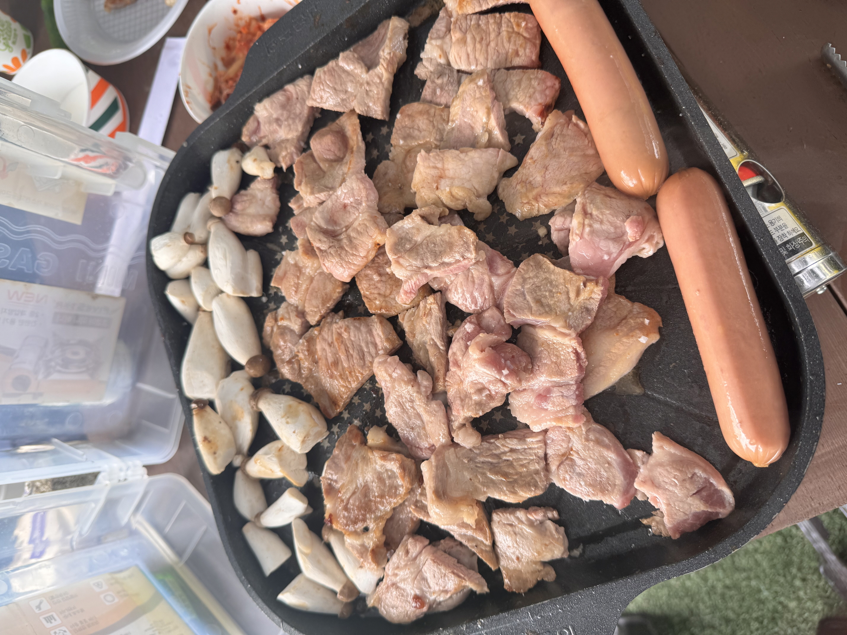 불판 위에 맛있게 구워지고 있는 돼지고기와 소세지, 새송이버섯이 찍힌 사진입니다.