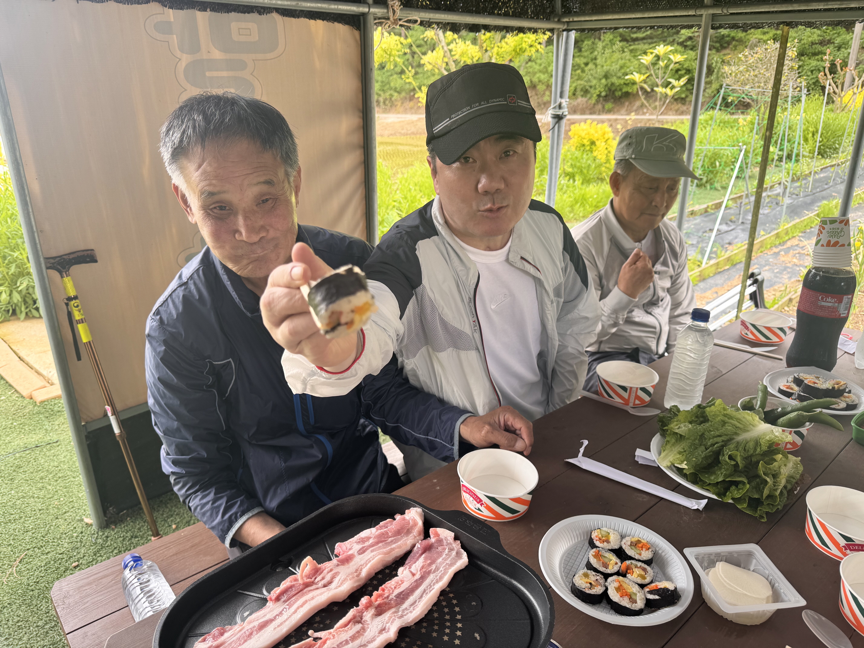 불판위에는 삼겹살이 구워지고 있고 재가장애인 한 분이 김밥을 집어 앞으로 내밀고 있는 사진입니다.