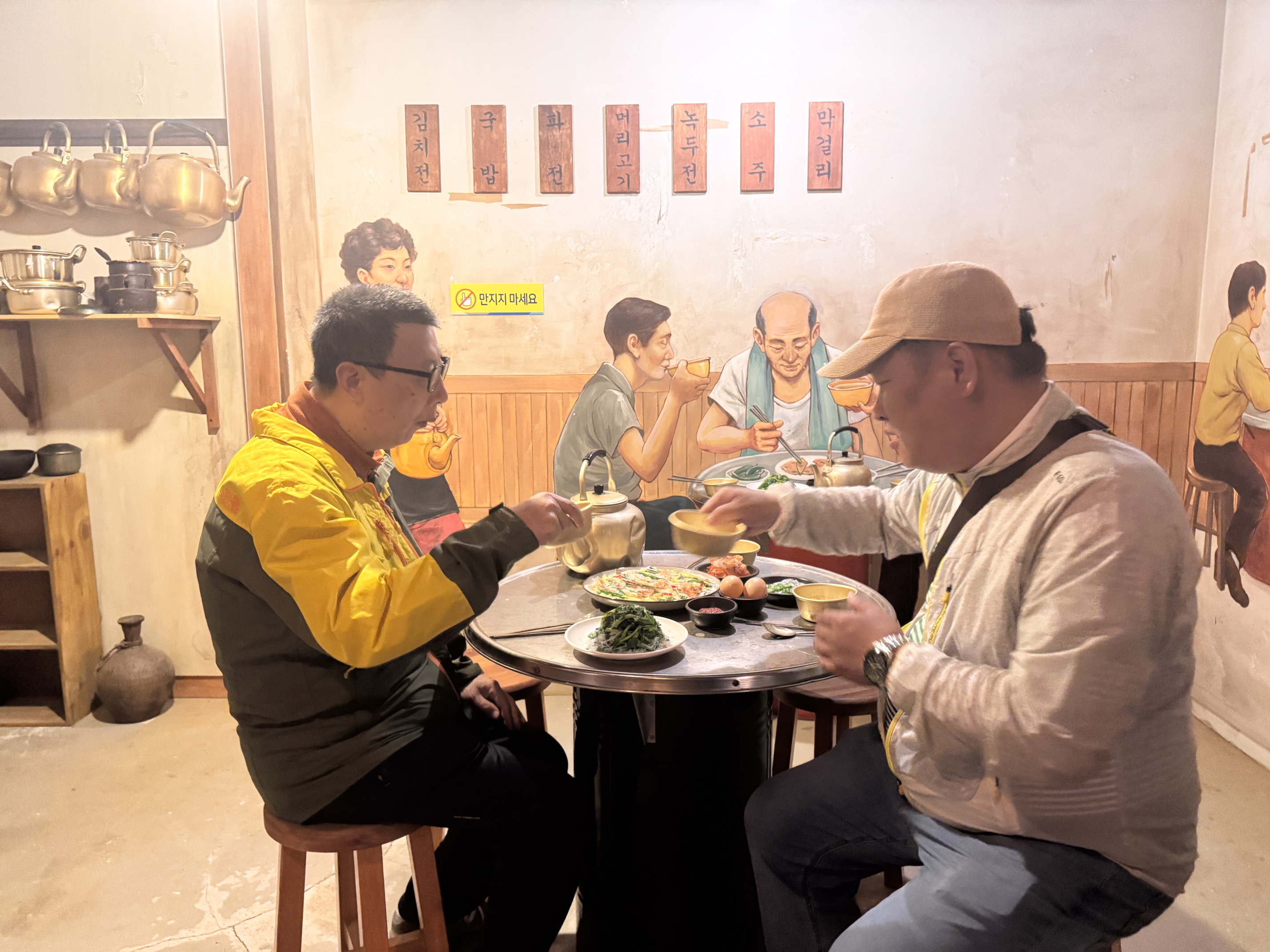 무안전통생활문화테마파크에 방문, 음식모형이 올려져 있는 테이블에서 막거리를 마시는 것처럼 흉내를 내고 있는 두명의 남성 재가장애인의 사진입니다.