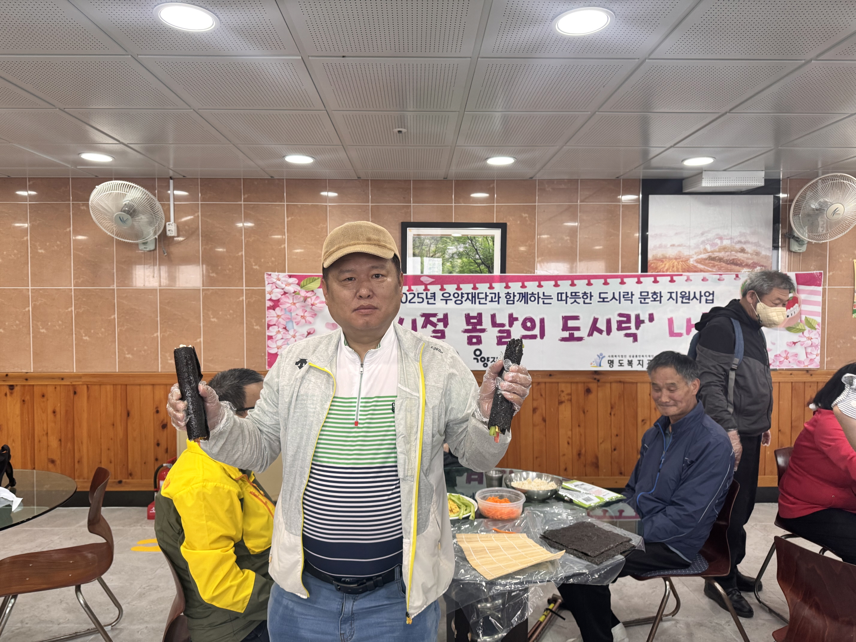 2025년 우양재단과 함께하는 따뜻한 도시락 문화지원사업. 그시절 봄날의 도시락 나들이라고 적힌 현수막 앞에서 남성 재가장애인 한분이 완성된 김밥을 들고 찍은 사진입니다.
