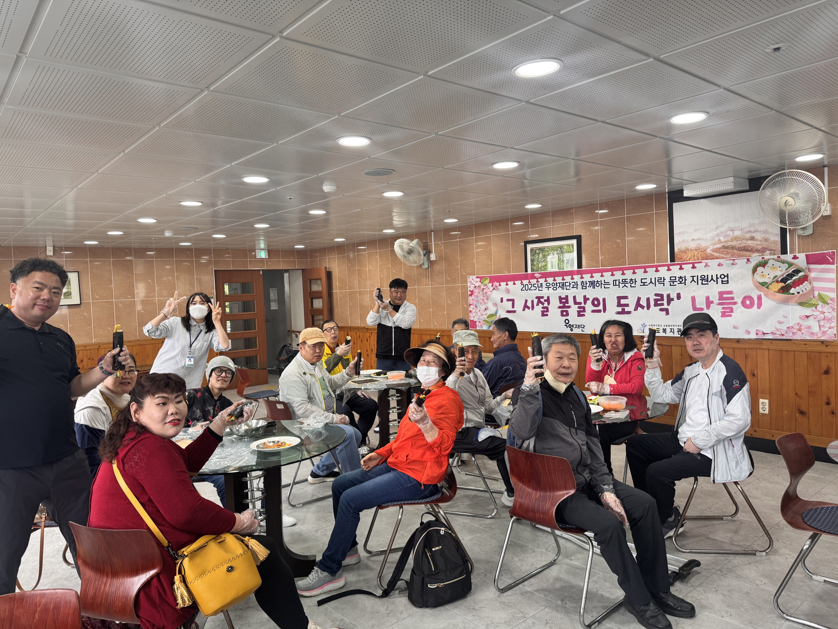 2025년 우양재단과 함께하는 따뜻한 도시락 문화지원사업. 그시절 봄날의 도시락 나들이라고 적힌 현수막이 관내 식당 벽면에 붙여 있고, 그 앞으로 김밥만들기를 함께한 재가장애인분들과 직원들이 모두 함께 자리에 앉아 완성된 김밥 한줄씩을 들고 찍은 단체사진입니다.