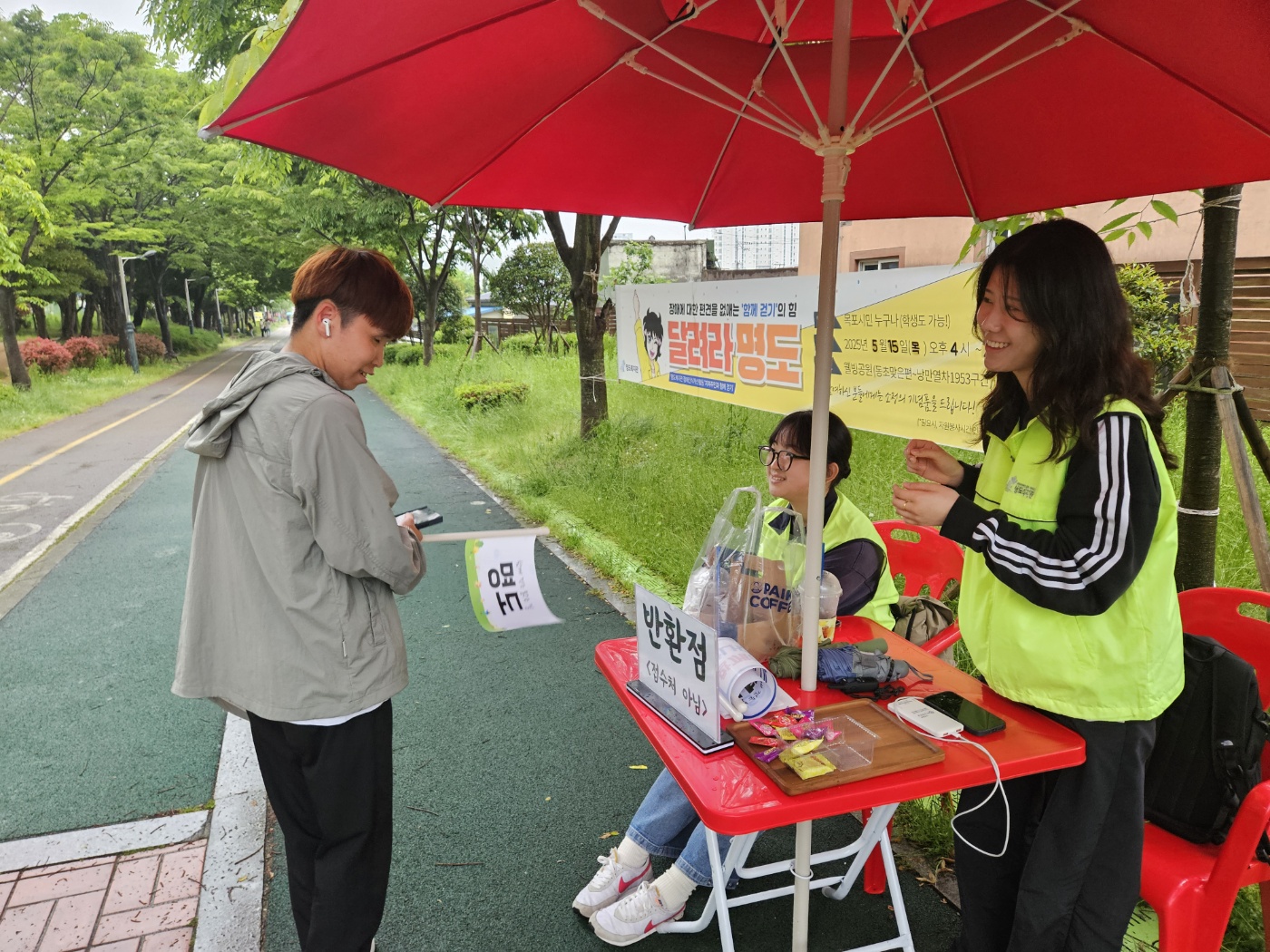 '명도'라고 적힌 깃발을 든 지역주민이 반환점에 들려 간식을 받으며 자원봉사자들과 이야기를 나누는 모습의 사진입니다. 