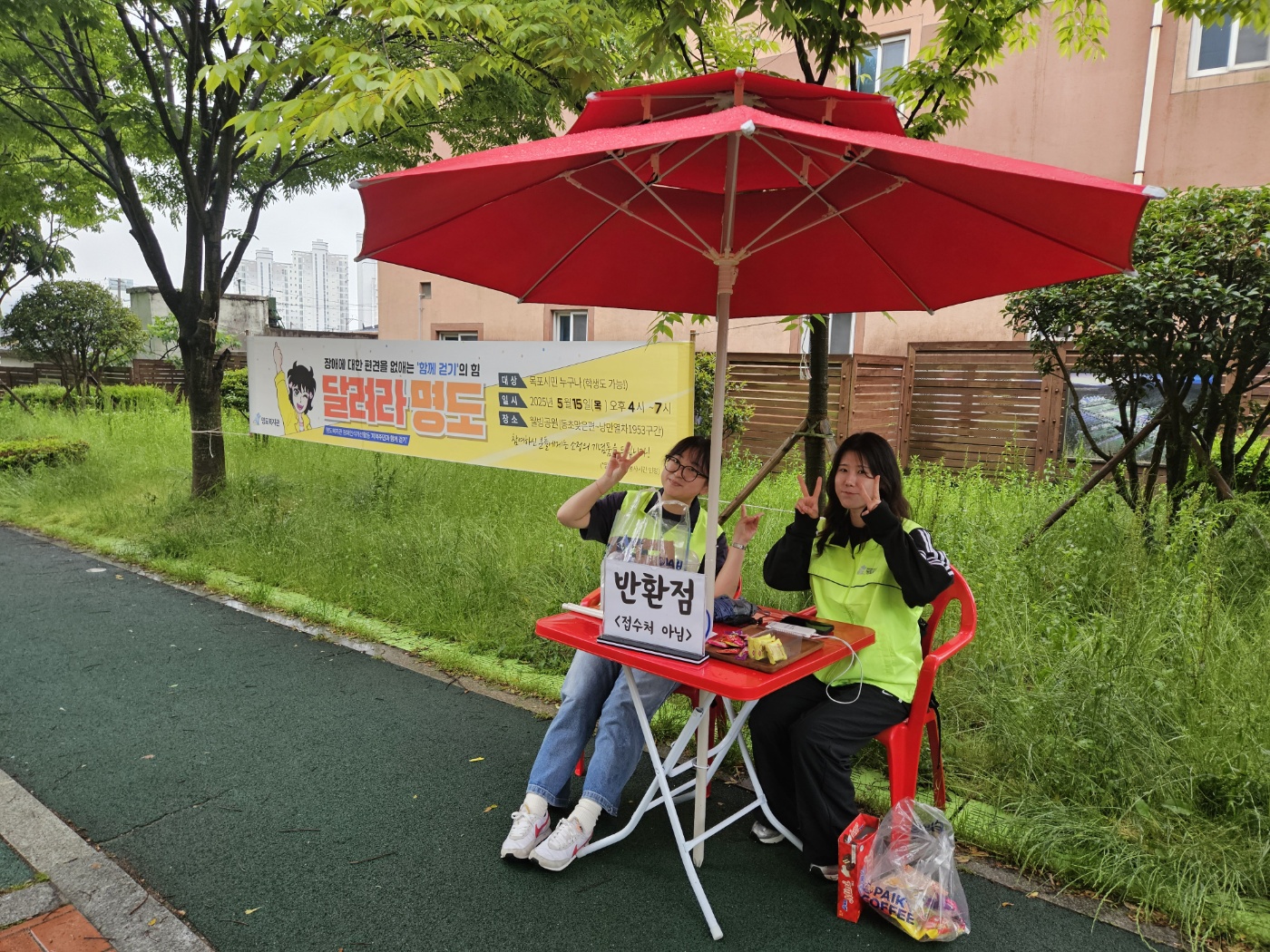 '달려라명도' 현수막 앞에 설치된 반환점 부스에서 브이포즈를 취하고 있는 두명의 자원봉사자 사진입니다. 