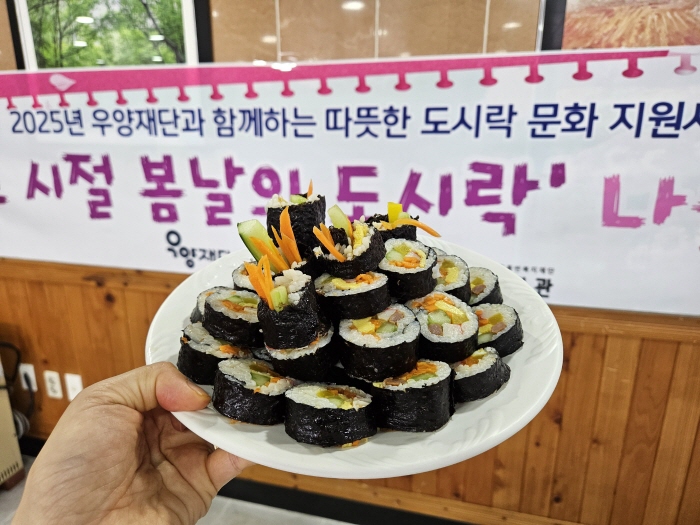 2025년 우양재단과 함께하는 따뜻한 도시락 문화지원사업. 그시절 봄날의 도시락 나들이라고 적힌 현수막을 배경으로 완성된 김밥을 잘라 접시에 담아 찍은 사진입니다.