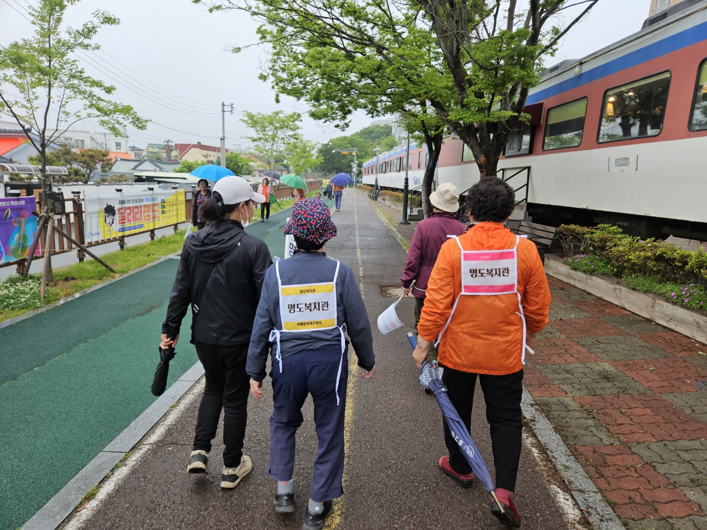 배번호띠를 메로 우산을 든 채로 함께걷기에 참여하고 있는 지역주민 3명의 뒷모습입니다. 뒷면의 배번호에는 명도복지관, 장애인식개선 이라고 적혀있습니다. 