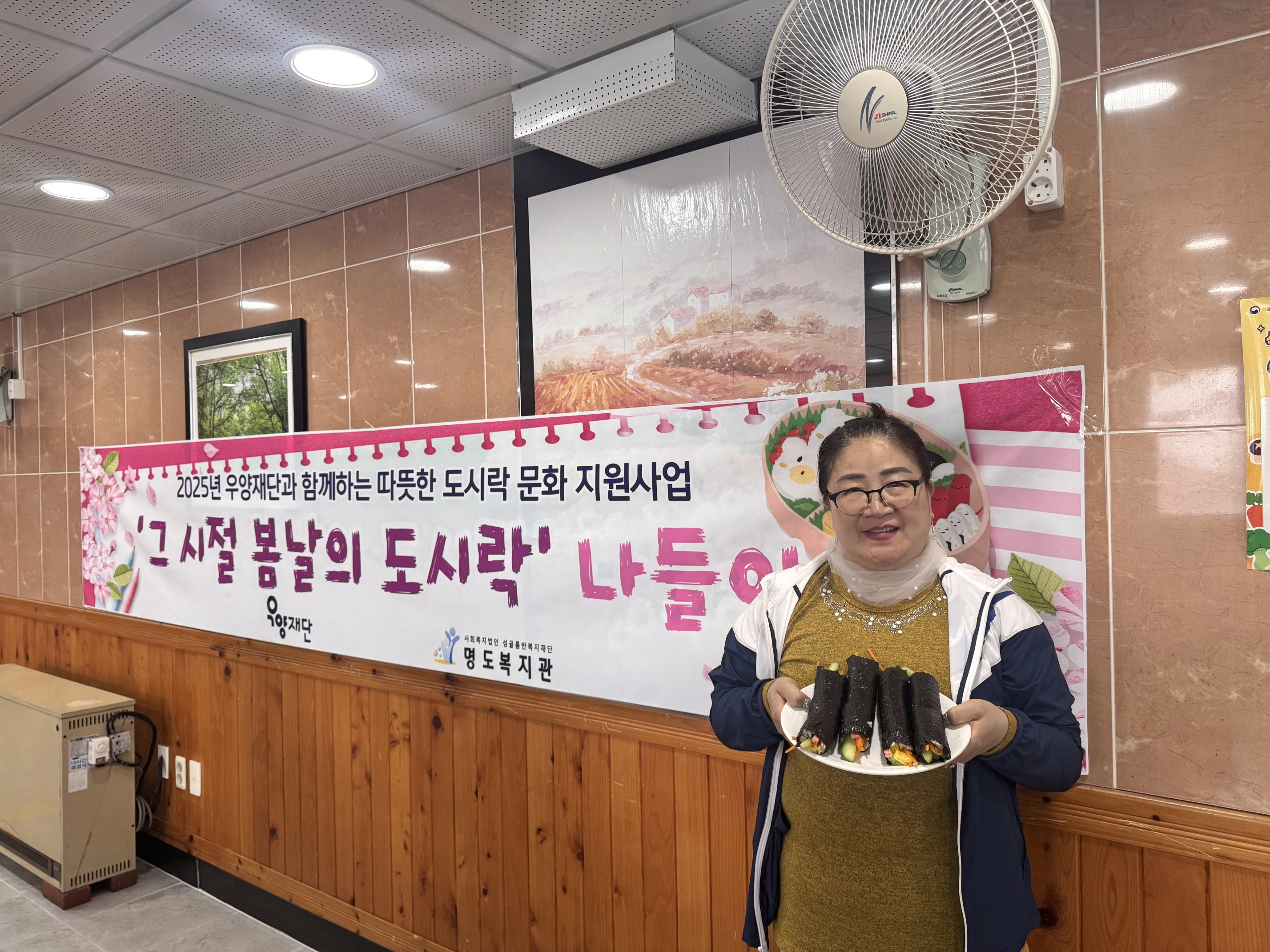 2025년 우양재단과 함께하는 따뜻한 도시락 문화지원사업. 그시절 봄날의 도시락 나들이라고 적힌 현수막 앞에서 완성된 김밥4줄을 접시에 담아 들고 찍은 여성 재가장애인분의 사진입니다.