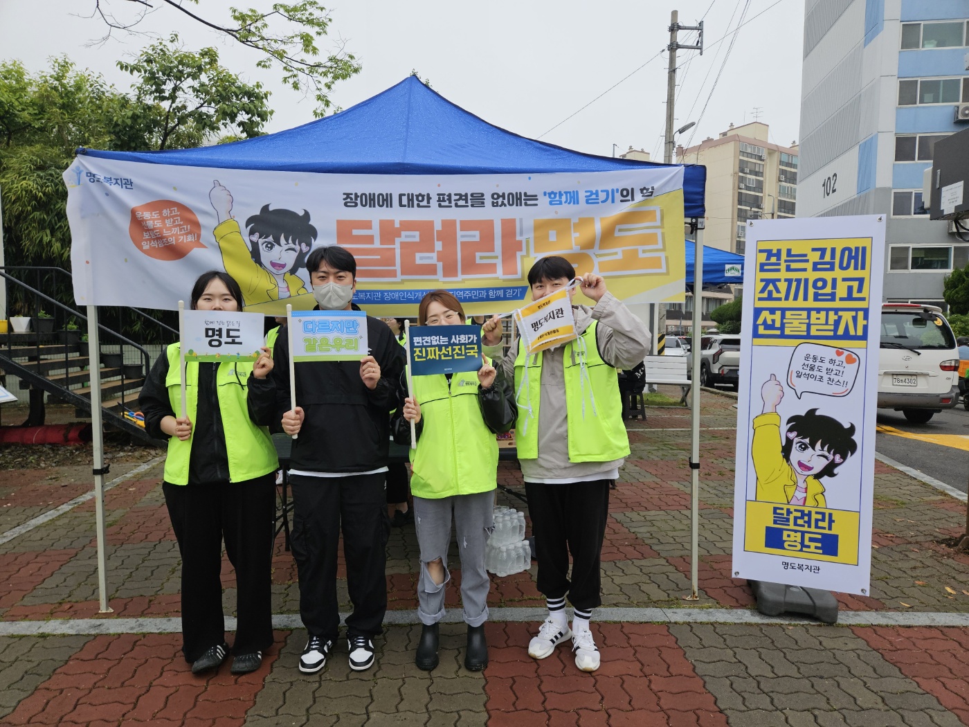 '달려라명도' 현수막이 걸린 천막앞에서 4명의 자원봉사자가 배번호와 깃발을 들고 찍은 사진입니다. 그 옆에는 '걷는김에 조끼입고 선물받자. 달려라명도'라고 적힌 X배너가 세워져 있습니다. 