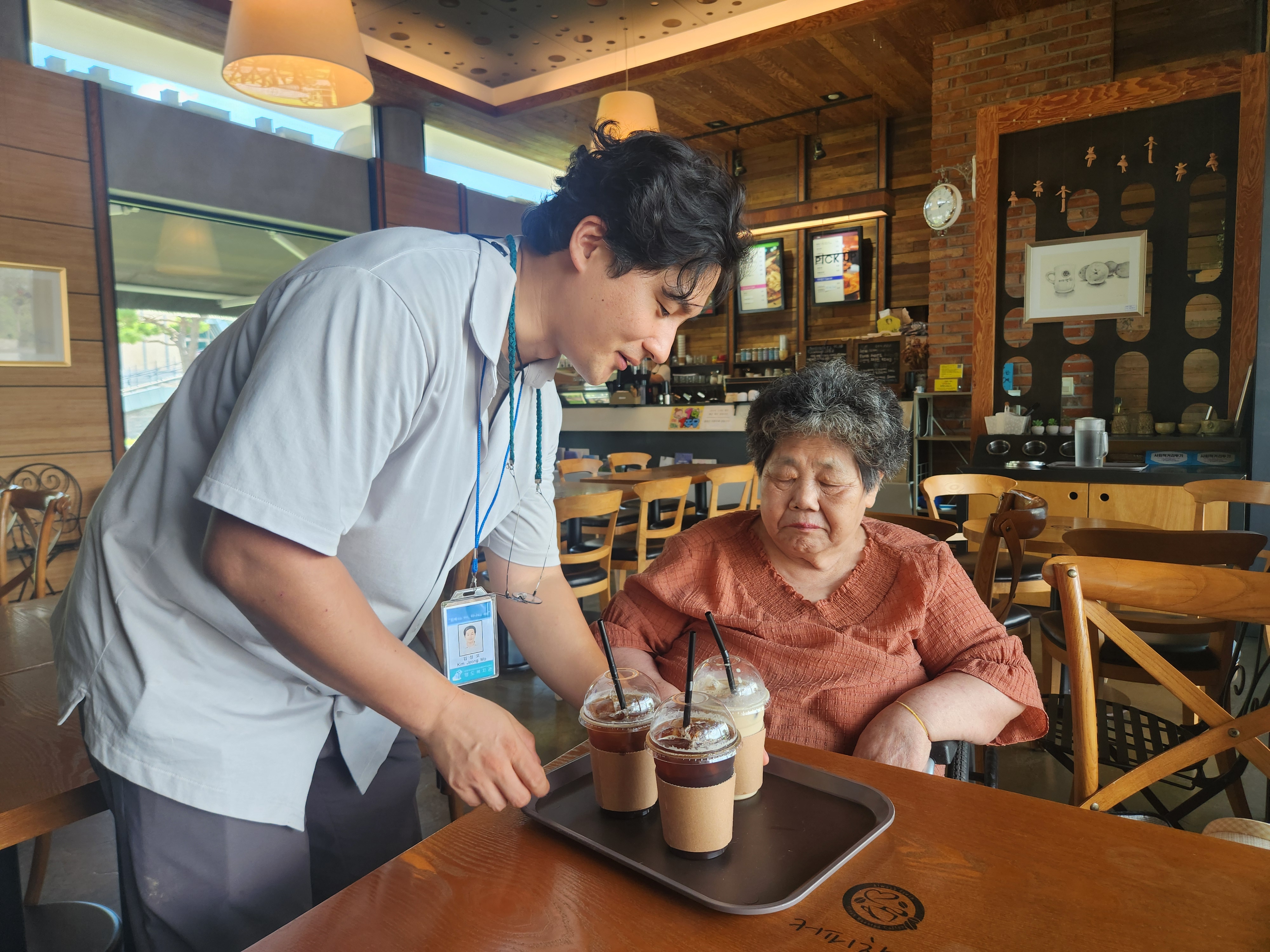 카페에서 이용인과 함께 음료를 먹기 위해 쟁반에 담긴 음료를 테이블로 옮기고 있는 담당 사회복지사의 모습입니다.