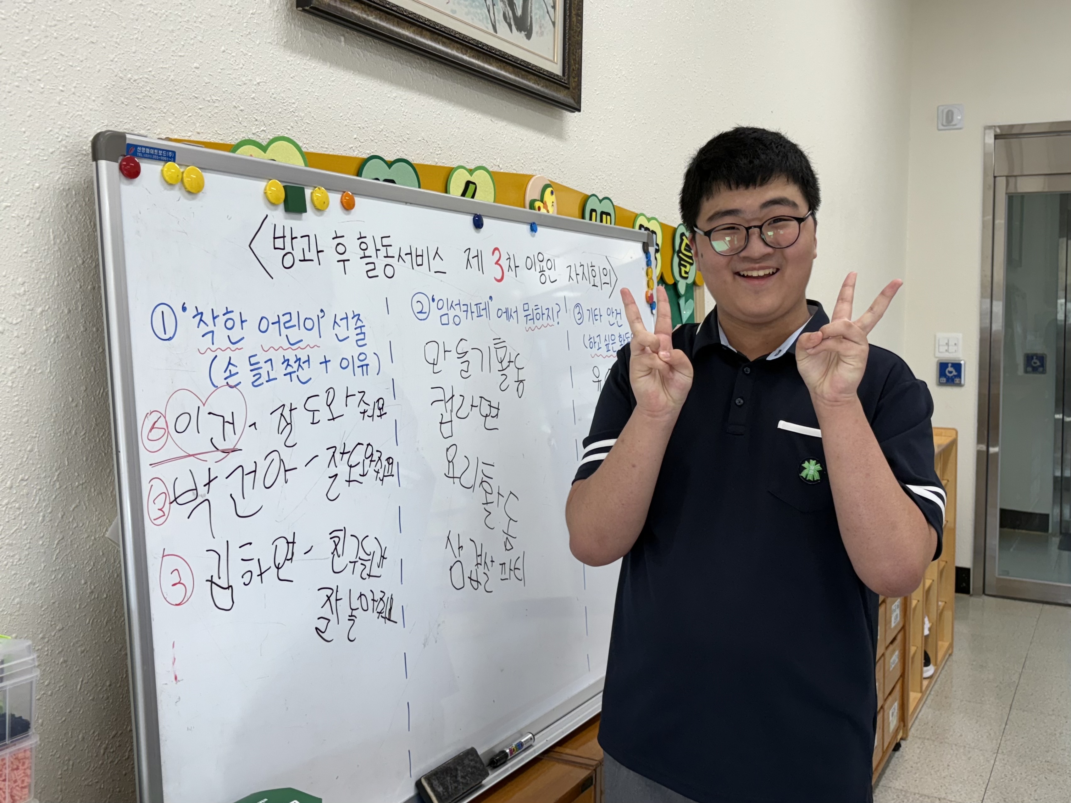 방과후교실의 회의 내용이 적힌 칠판앞에서 사회자 역할을 맡은 이용인이 포즈를 취하며 밝게 웃고 있는 모습입니다.