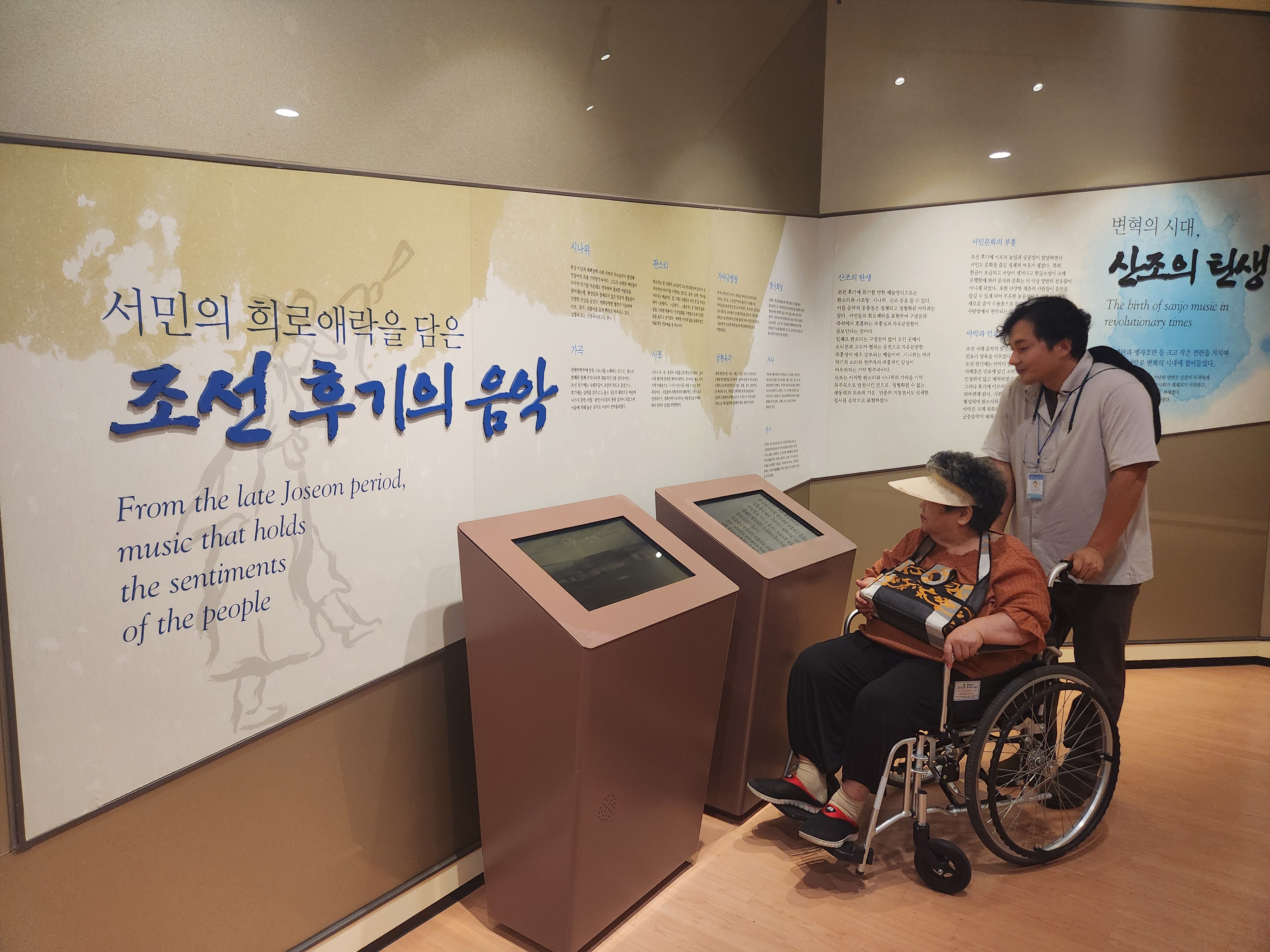 휠체어에 탄 이용인 담당 사회복지사가 함께 트로트가요센터를 관람하고 있는 모습입니다.