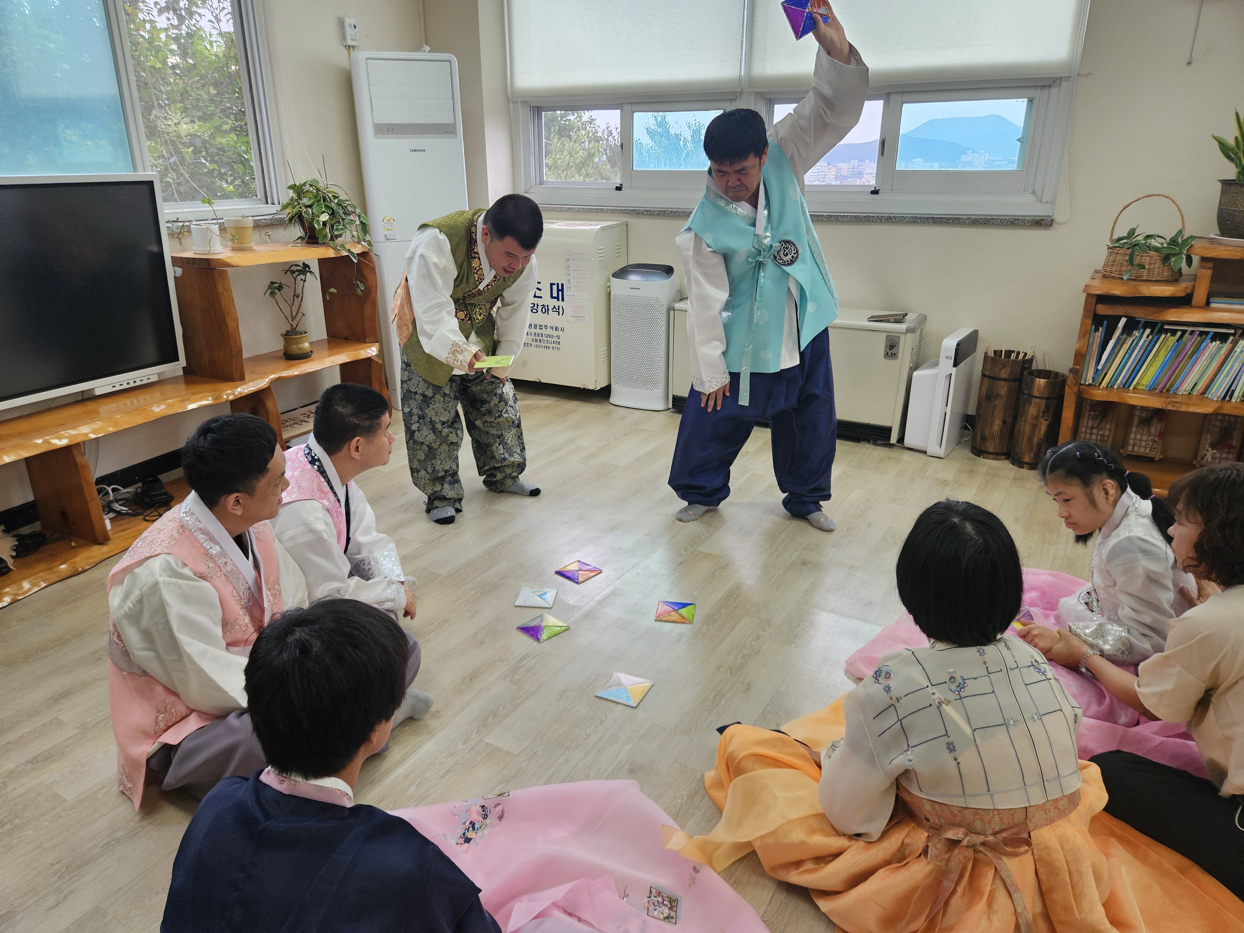 한복을 차려입은 이용인들이 동그랗게 둘러 앉아있고, 그 가운데에 남성 이용인 2명이 서서 딱지를 치고 있는 사진입니다.
