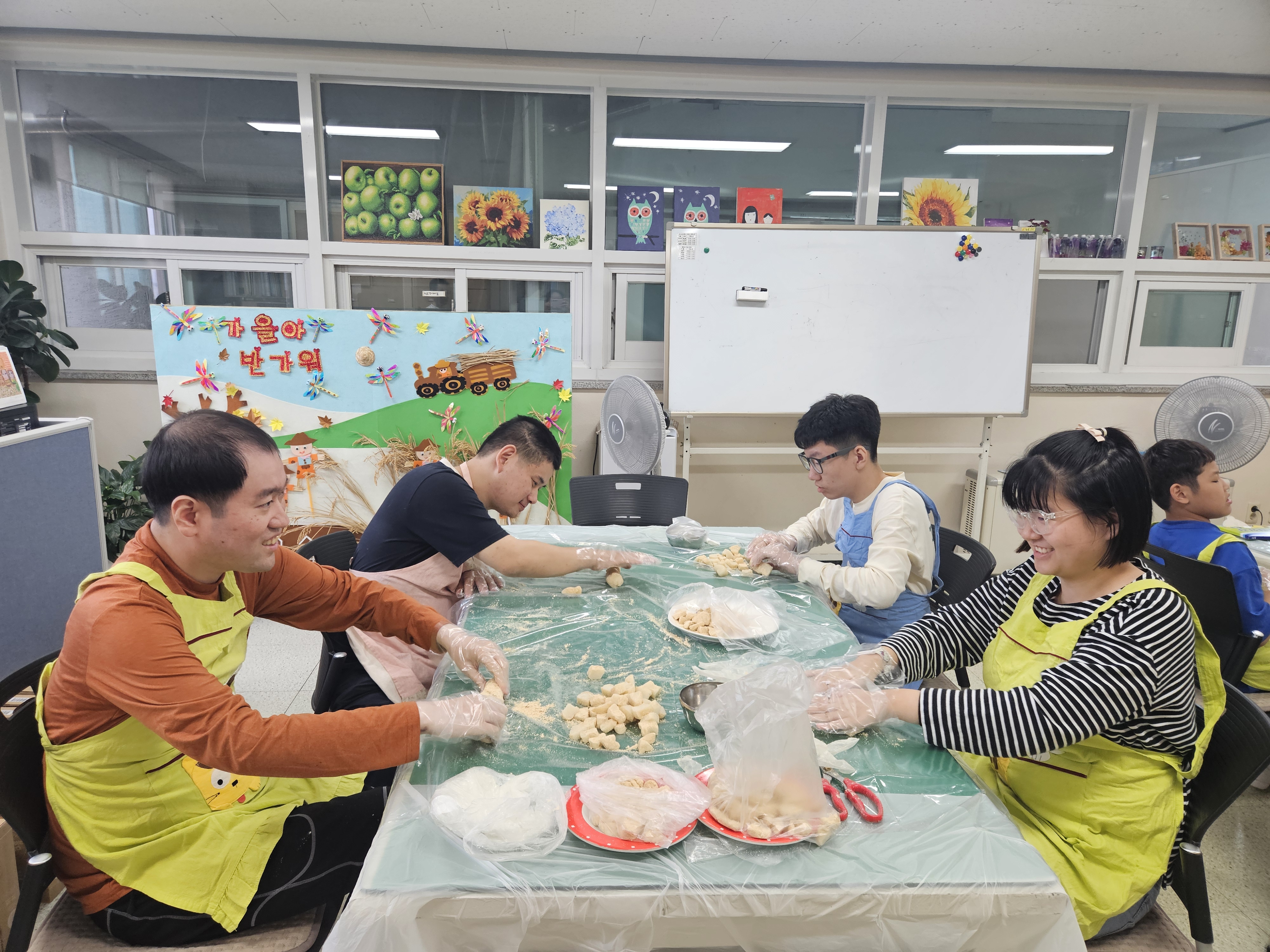 이용인들이 앞치마를 한 채로 책상에 앉아 인절미 떡 반죽에 콩가루를 묻히고 있는 사진입니다.