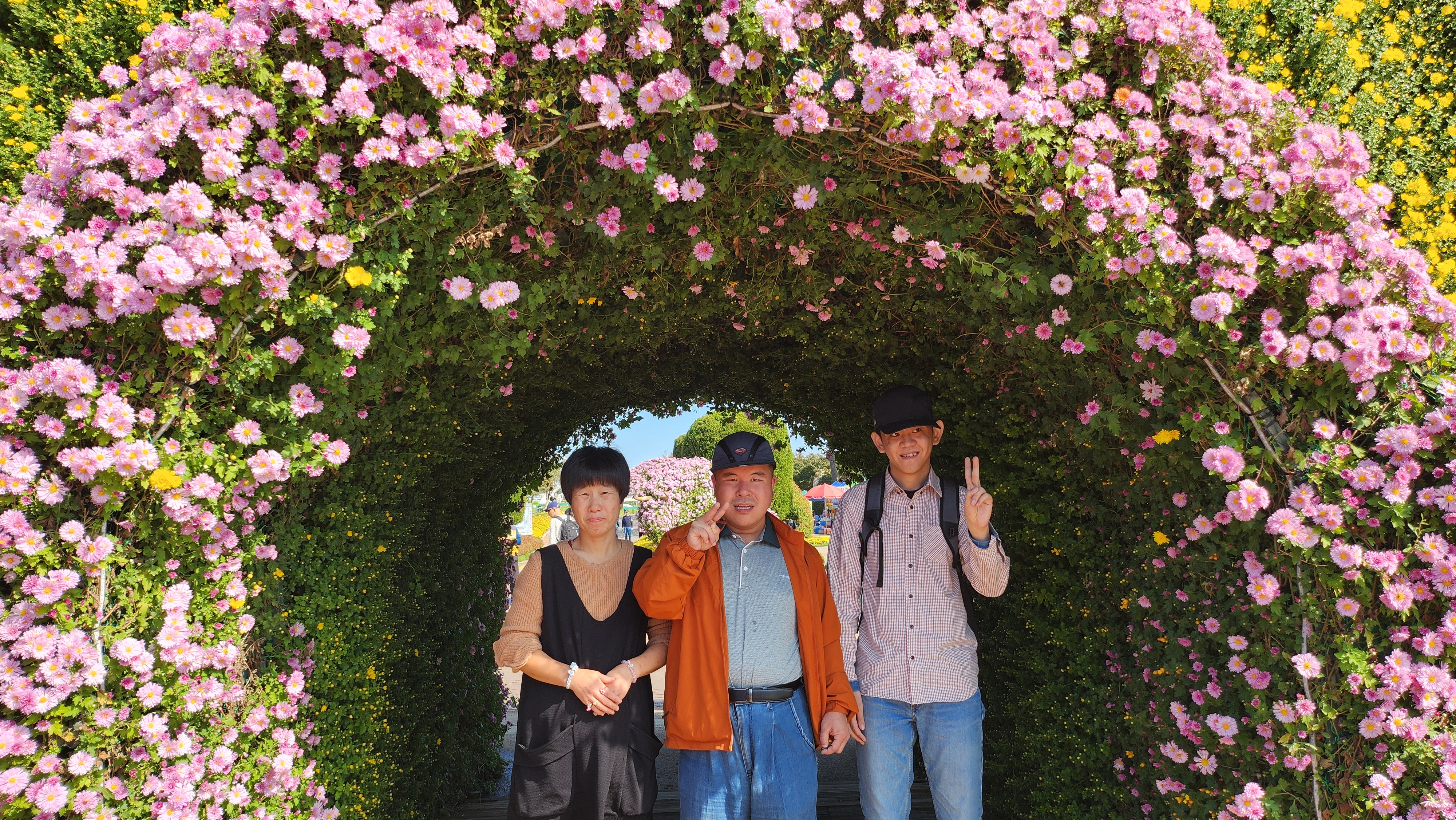 분홍색 꽃 터널 가운데에 여성 이용인 1명, 남성 이용인 2명이 서서 '브이'하고 포즈를 취하고 있는 모습입니다.